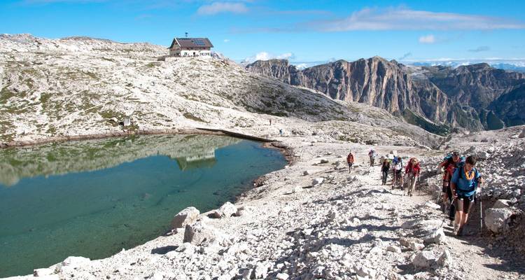 Hikers walking towards a mountain hut with rugged terrain and a small lake.