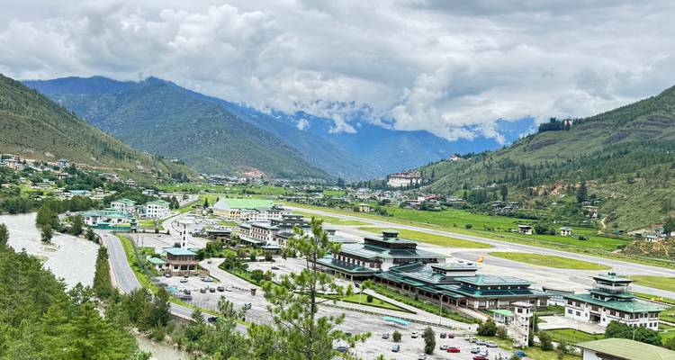 Vue aérienne de la piste de l'aéroport de Paro flanquée d'une rivière et de montagnes sous des nuages fragmentés.
