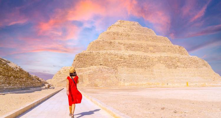 Femme vêtue d'une robe rouge éclatante marchant vers la Pyramide à Degrés de Djéser sous un ciel de coucher de soleil rose dramatique