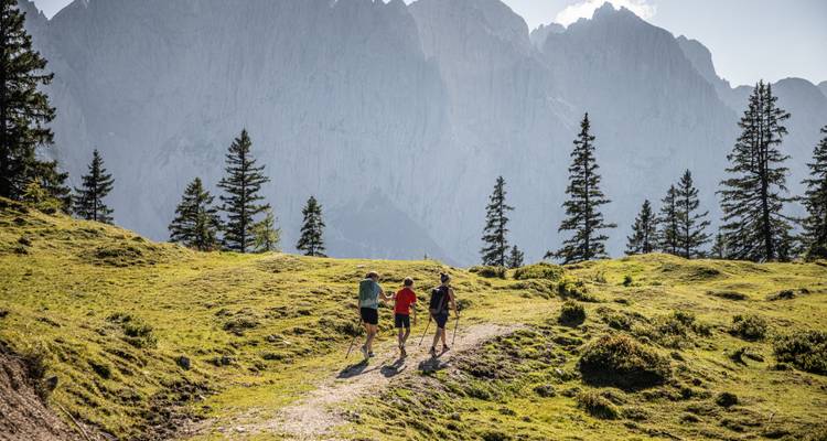 Tres personas caminando por un sendero con hierba con montañas de fondo.