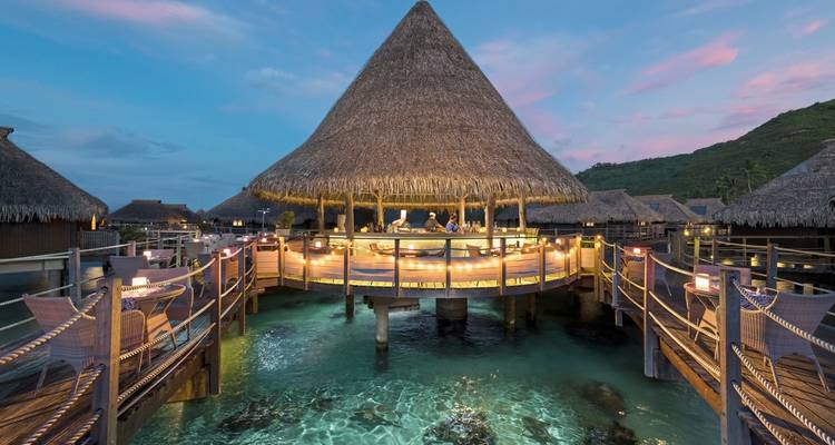 Coin repas sur pilotis au-dessus d'une eau claire au coucher du soleil.