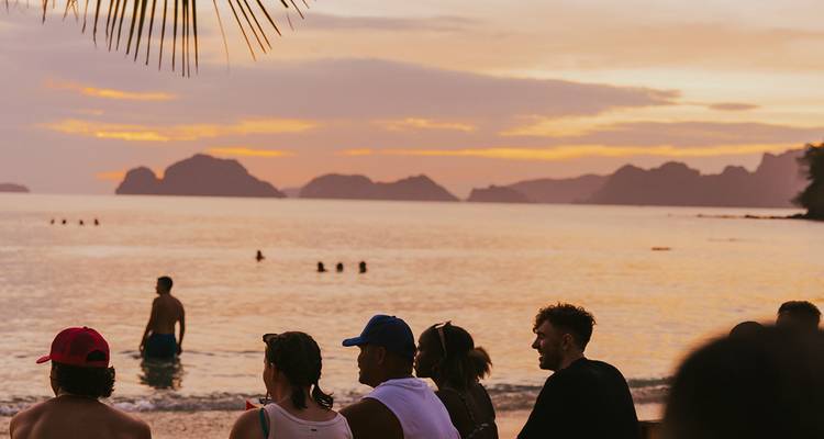 Los viajeros se sientan en una playa tropical observando un atardecer naranja sobre islas de piedra caliza.