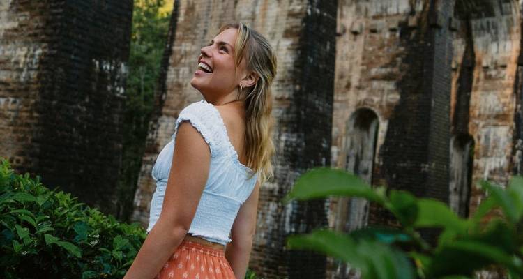 Viajero sonriente con una blusa blanca parado junto a pilares de ladrillo antiguos rodeado de exuberante vegetación en Sri Lanka