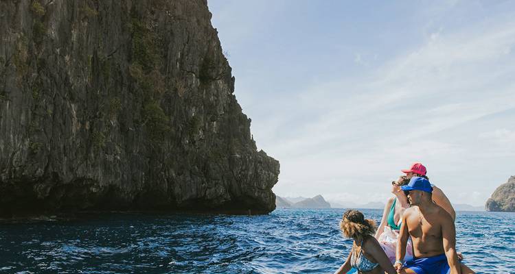 Amigos en un bote señalan hacia imponentes acantilados de piedra caliza en aguas azules cristalinas.