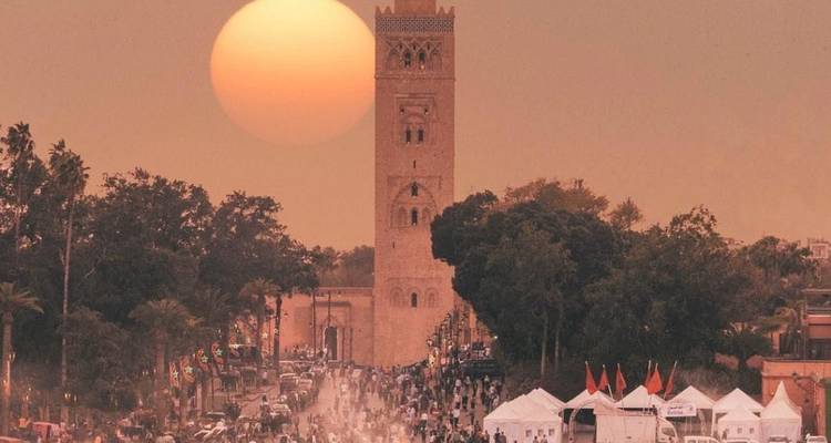 Calle del mercado con una torre de mezquita y sol poniente en Marrakech.