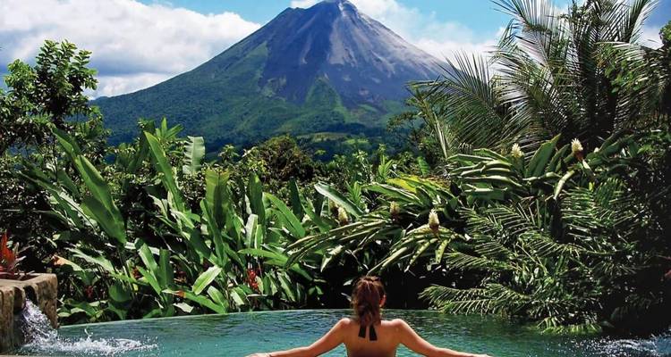 Personne dans une piscine avec vue sur un volcan, entourée de verdure.