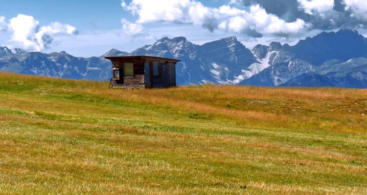 Cabane solitaire avec de vastes champs herbeux et chaîne de montagnes.