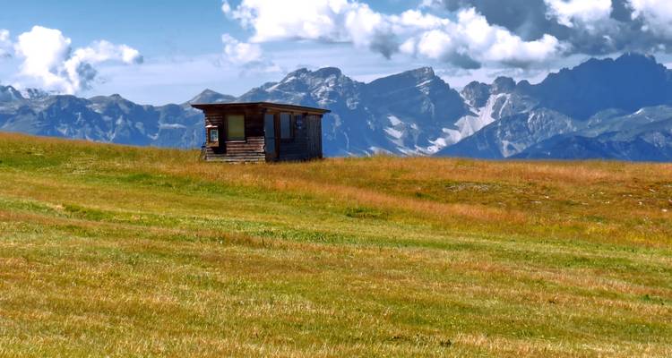 Eine einsame Holzhütte sitzt auf welligem alpinem Grünland unter einer dramatischen Dolomiten-Silhouette und flauschigen Wolken.