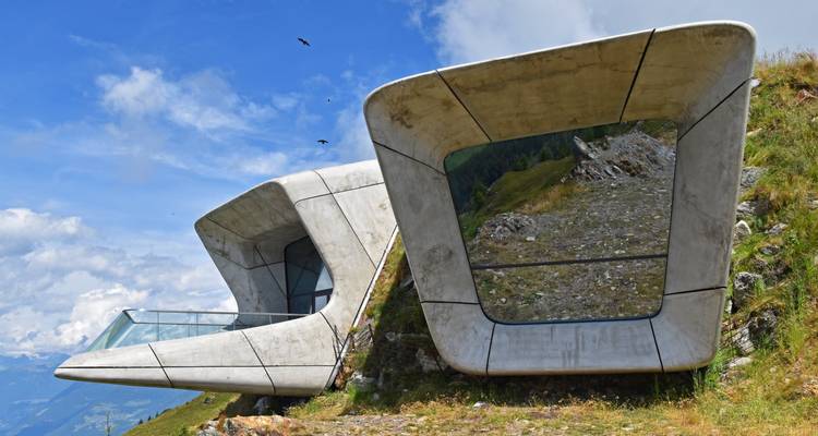 Die futuristische Betonkonstruktion des Messner Mountain Museum ragt aus einem grasbewachsenen Hang vor alpinen Ausblicken auf.