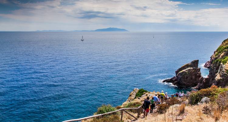 Senderistas descendiendo por un sendero de madera a lo largo de un acantilado mediterráneo escarpado hacia el mar azul profundo.