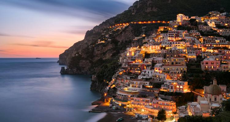 Vista crepuscular de las casas en la ladera de Positano brillando con luces sobre un mar en calma.