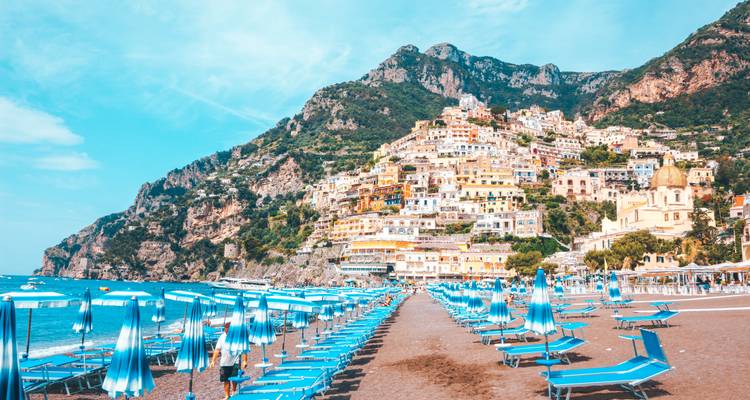 Escena de playa luminosa con filas de tumbonas azules de cara al colorido pueblo costero de Positano en el acantilado.