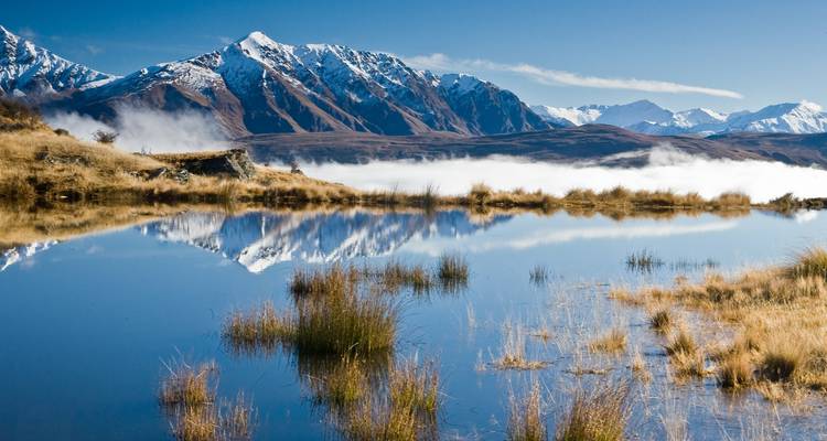Montañas cubiertas de nieve reflejándose en un lago sereno con hierba dorada.