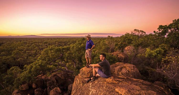 Two people standing on rocks, enjoying a view under a pink sunset.