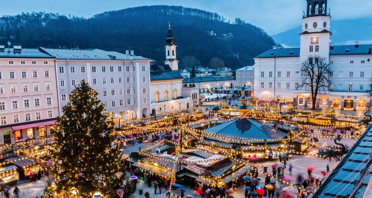 Marché de Noël festif sur une place de ville avec un grand sapin décoré et des guirlandes lumineuses scintillantes au crépuscule.