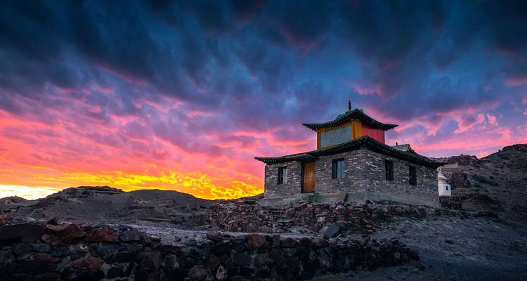 Geïsoleerde stenen tempel tegen een levendige vurige zonsonderganglucht boven een rotsachtig landschap.