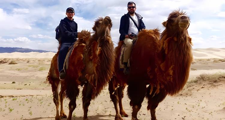 Twee reizigers rijden op ruige Bactrische kamelen over zandduinen onder een heldere Mongoolse hemel.