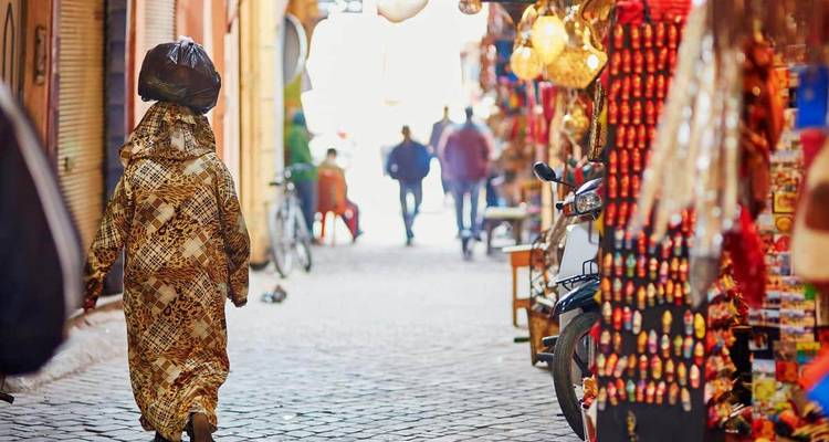 Une rue étroite dans un marché marocain avec des gens qui font leurs courses et des marchandises colorées exposées des deux côtés.
