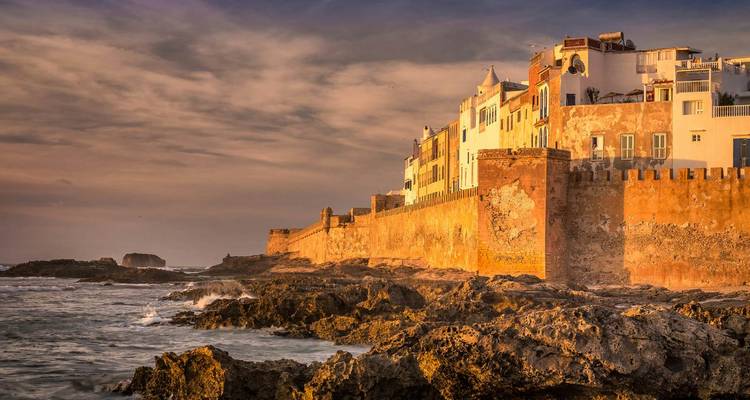 Une vue côtière d'Essaouira avec des vagues qui se brisent contre les falaises rocheuses et les bâtiments de la vieille ville au coucher du soleil.