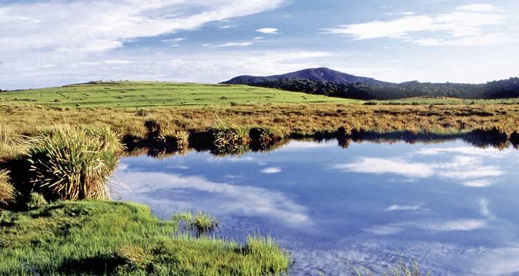 Serene lake reflecting the sky with grassy banks and distant mountains.