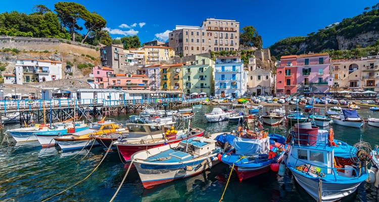 Colorful fishing boats moored in a picturesque harbour lined with pastel houses on a sunny day.