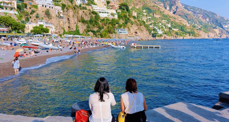 Two women sit on waterfront steps looking out at a lively Positano beach and the azure sea.