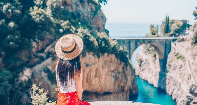 Young woman in a straw hat admires a dramatic stone bridge spanning a turquoise gorge along the Amalfi Coast.