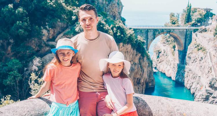 Father with two daughters posing happily at a scenic stone bridge viewpoint over a coastal gorge.