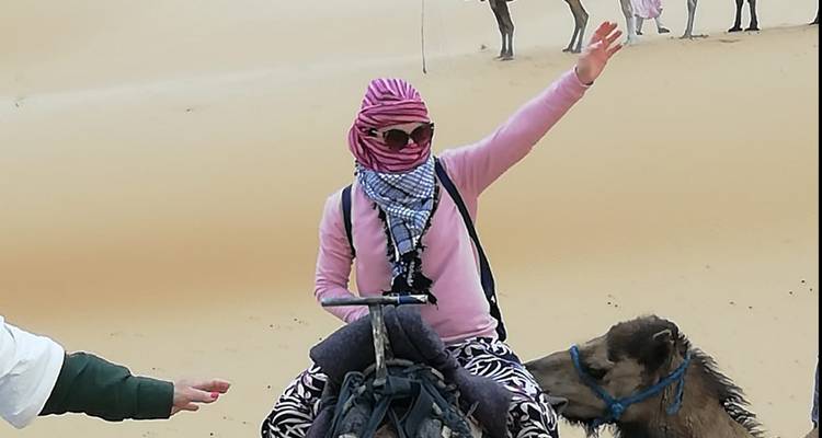 Reisender mit Kopftuch und Sonnenbrille winkt, während er auf einem Kamel auf weichem Wüstensand sitzt