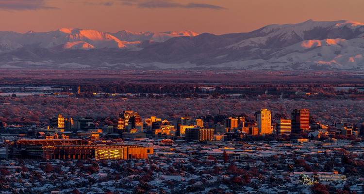 Un paysage urbain de Christchurch au coucher du soleil, avec des montagnes enneigées en arrière-plan, créant un contraste saisissant entre les couleurs chaudes et froides.