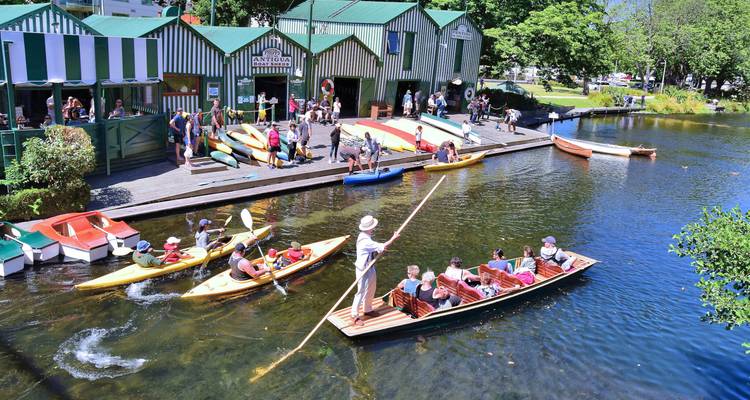 Une scène animée avec des gens naviguant sur une rivière à côté de hangars à bateaux peints aux couleurs vives, entourés d'une verdure luxuriante.