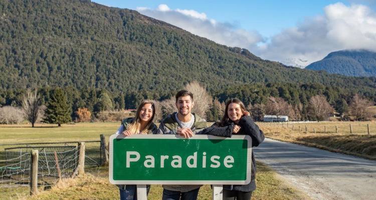 Trois personnes tenant un panneau 'Paradise' avec des montagnes en arrière-plan.