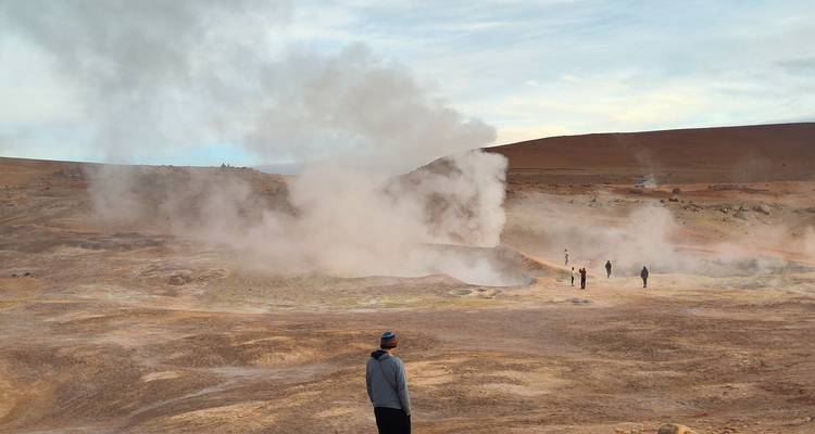 Mensen die wandelen tussen stoomgeisers in een kaal landschap.