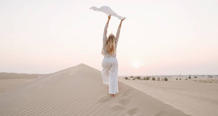 Personne debout sur une dune de sable tenant un tissu, face au lever du soleil