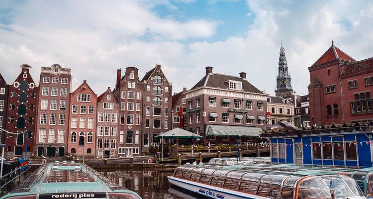 Tall, narrow canal houses beside tour boat docks under a partly cloudy Amsterdam sky.