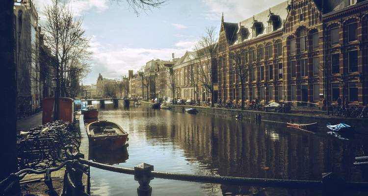 Quiet Amsterdam canal lined with bikes, moored boats and elegant brick buildings on an overcast day.