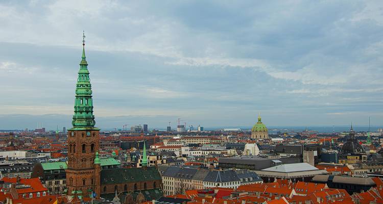 Wide city panorama with tall green church spire and domed marble cathedral beneath a grey sky.