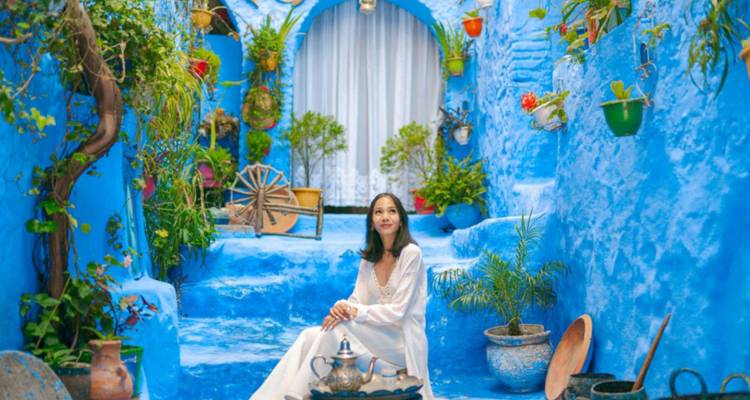 Une femme en robe blanche fluide est assise dans une cour d'un bleu vif décorée de poteries marocaines et de plantes à Chefchaouen.