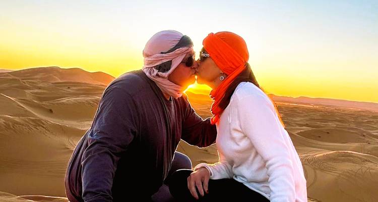 Un couple portant des foulards du désert s'embrasse au sommet de dunes de sable au coucher du soleil.