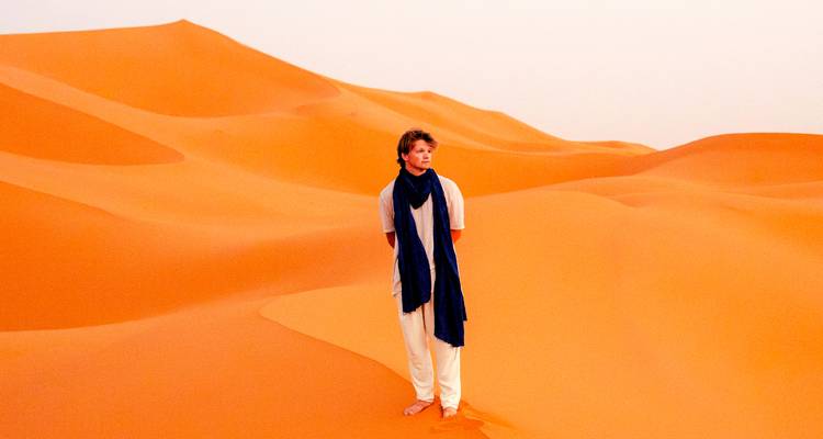 Un voyageur solitaire se tient pieds nus sur des dunes de désert orange saisissantes sous un ciel pâle.