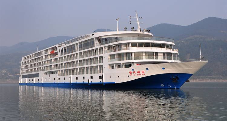 Moderno crucero blanco anclado en un río tranquilo con montañas brumosas elevándose de fondo.