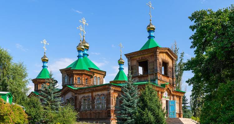 Orthodoxe Holzkirche mit bunten Dächern und goldenen Kuppeln, umgeben von Bäumen.