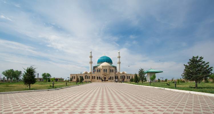 Vista amplia de una mezquita moderna con cúpula turquesa y dos minaretes gemelos al final de una plaza con patrón decorativo.