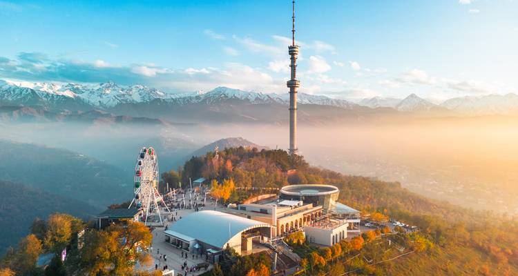Vista aérea al atardecer sobre la colina Kok-Tobe de Almaty con la torre de televisión, la rueda de la fortuna y las montañas nevadas más allá.
