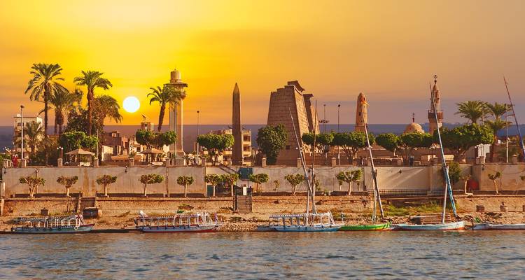 Waterfront view with ancient structures and sailing boats at sunset.