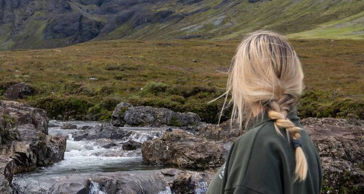 Una persona con cabello rubio largo observa un arroyo rocoso en un paisaje montañoso.