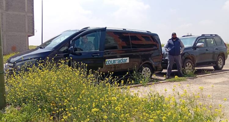 Ein schwarzer Personenvan mit der Aufschrift einer Tourismusfirma parkt neben gelben Wildblumen, während zwei Fahrer in der Nähe stehen.