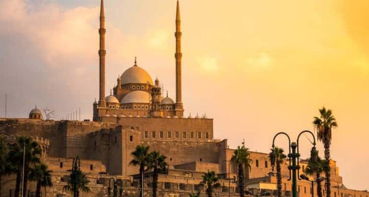 Die Moschee von Muhammad Ali mit Zwillingsminaretten, die im warmen Sonnenuntergangslicht über den Mauern der Zitadelle von Kairo und palmengesäumten Straßenlaternen leuchten.