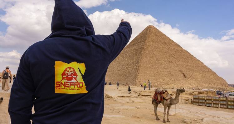 Persona señalando una pirámide con un camello cerca.