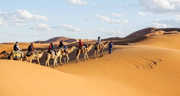 Chameaux en file avec des cavaliers traversant des dunes de sable.